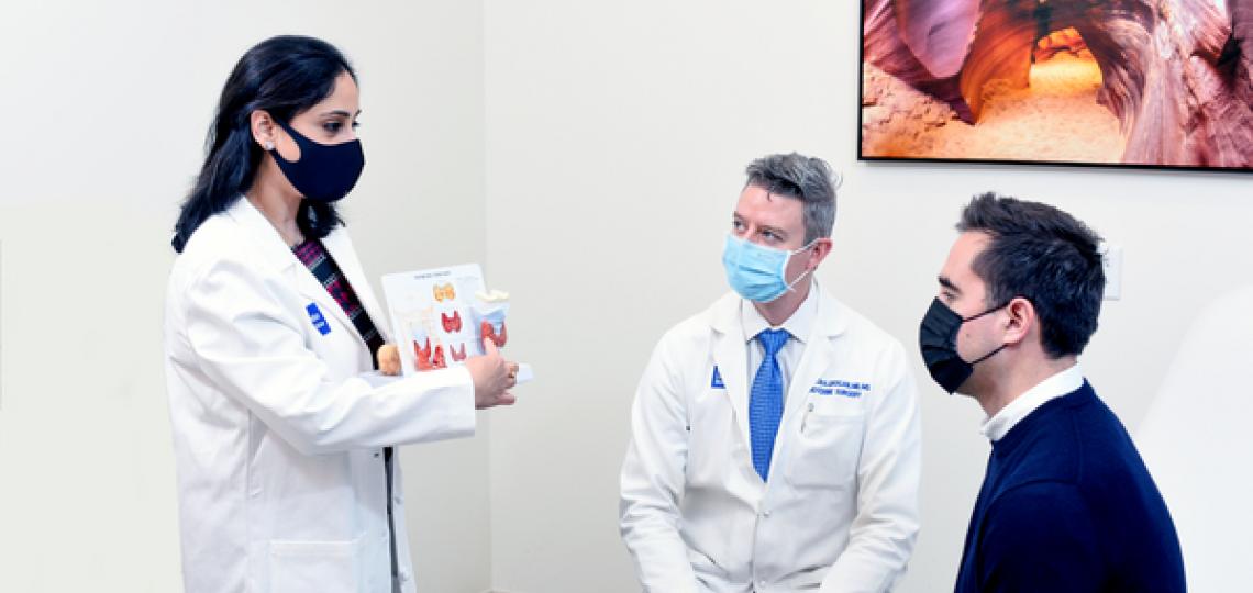 A masked female doctor explaining a diagram of the thyroid and parathyroid to a masked male doctor and masked male patient.