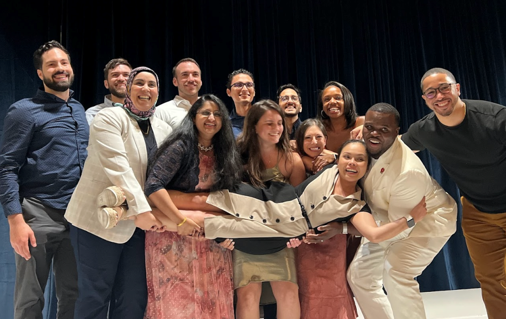 Emergency Medicine Residency Wellness members gather during a group meeting. They stand together in a row, with the group holding one person aloft in a horiztonal reclining position.