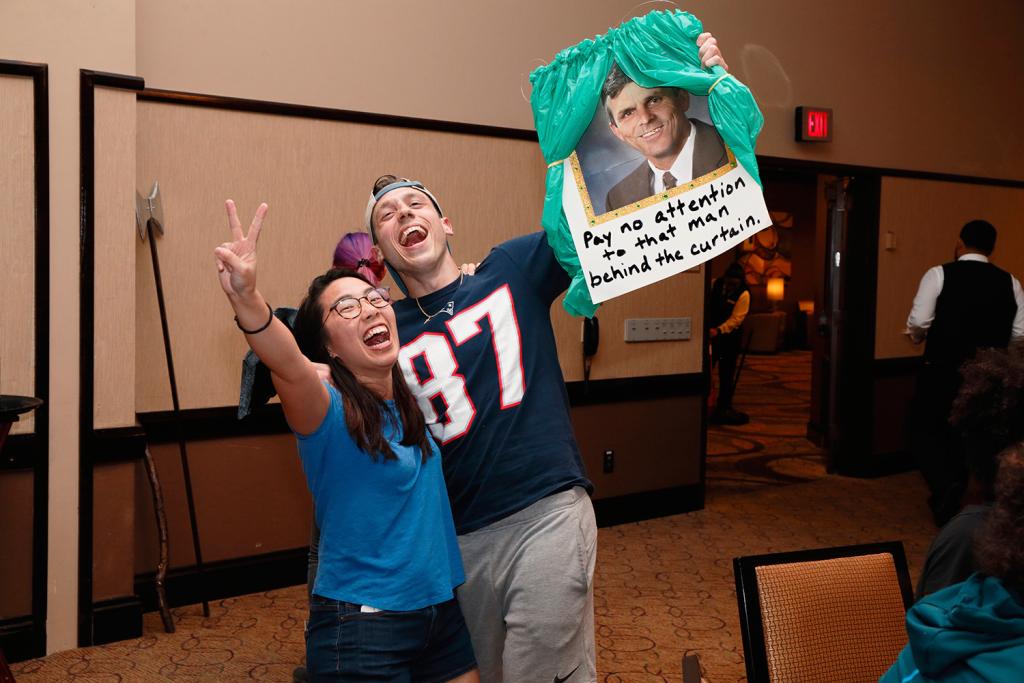 Two people cheering. One flashes a peace sign, the other holds a photo of a man with the words "Pay no attention to that man behind the curtain."