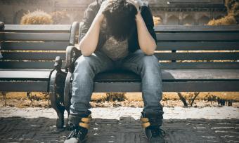 Photo of a man sitting on a bench holding his head in his hands. 