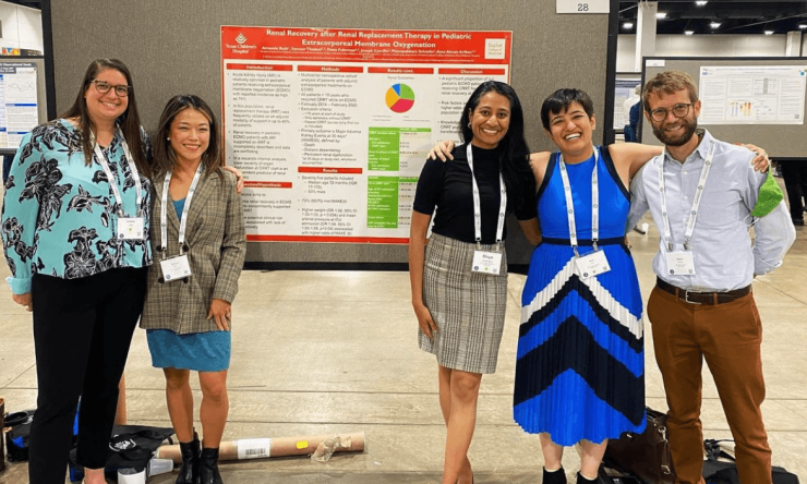 Five Pediatric Critical Care Medicine fellows during a research poster presentation. The group is posed on the sides of a poster with their arms on each other's shoulders.