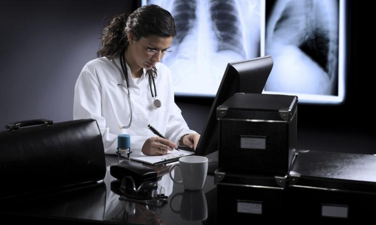 A female doctor taking notes and looking at a computer while a chest X-ray of the lungs displays on the wall.