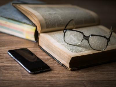 Photo of an open book with reading glasses sitting on the open pages and a cell phone next to the book. 
