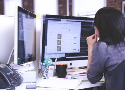 Photo taken from behind of a woman sitting at her desk computer hunched over  looking worried.