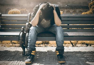 Photo of a man sitting on a bench holding his head in his hands. 