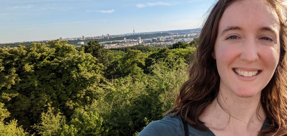 Katherine Pendleton stands on a hill overlooking a town. The view is a selfie taken with Pendleton facing the camera followed by a long line of trees and a city behind it.