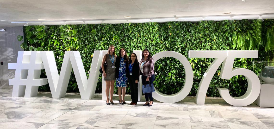 Four people standing in front of a large sign that reads #WHO75