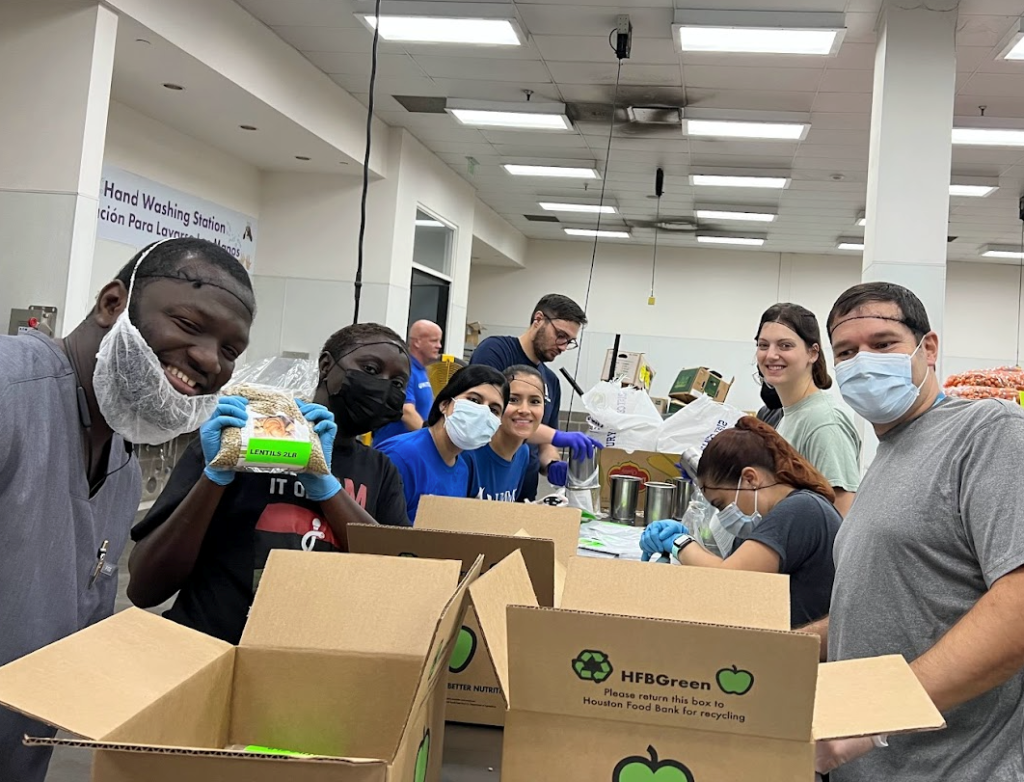 Emergency Medicine Residency Wellness members volunteer at the Houston Food Bank. The group is wearing masks and hair nets while packing food into boxes.