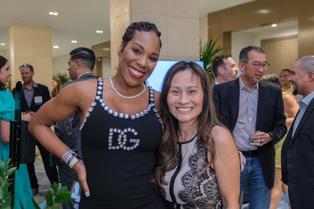 Two people smiling for the camera with a large group of laughing people behind them
