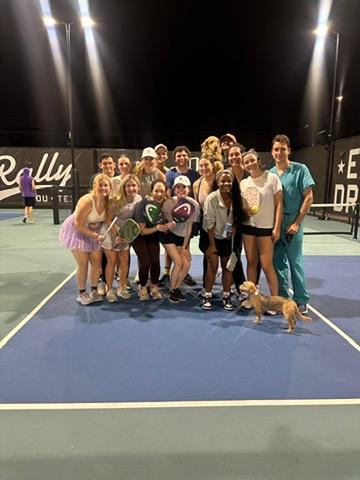 A large group of smiling people posing on a Pickleball court at night, along with a little dog.