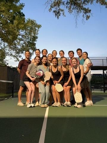 A large group of smiling people posing on a Pickleball court on a sunny day.