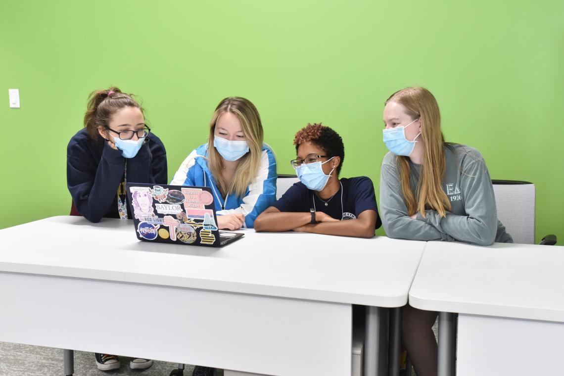 Four students in face masks huddle together behind a laptop