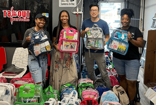 Volunteers holding bookbags full of supplies while standing amidst dozens more bags.