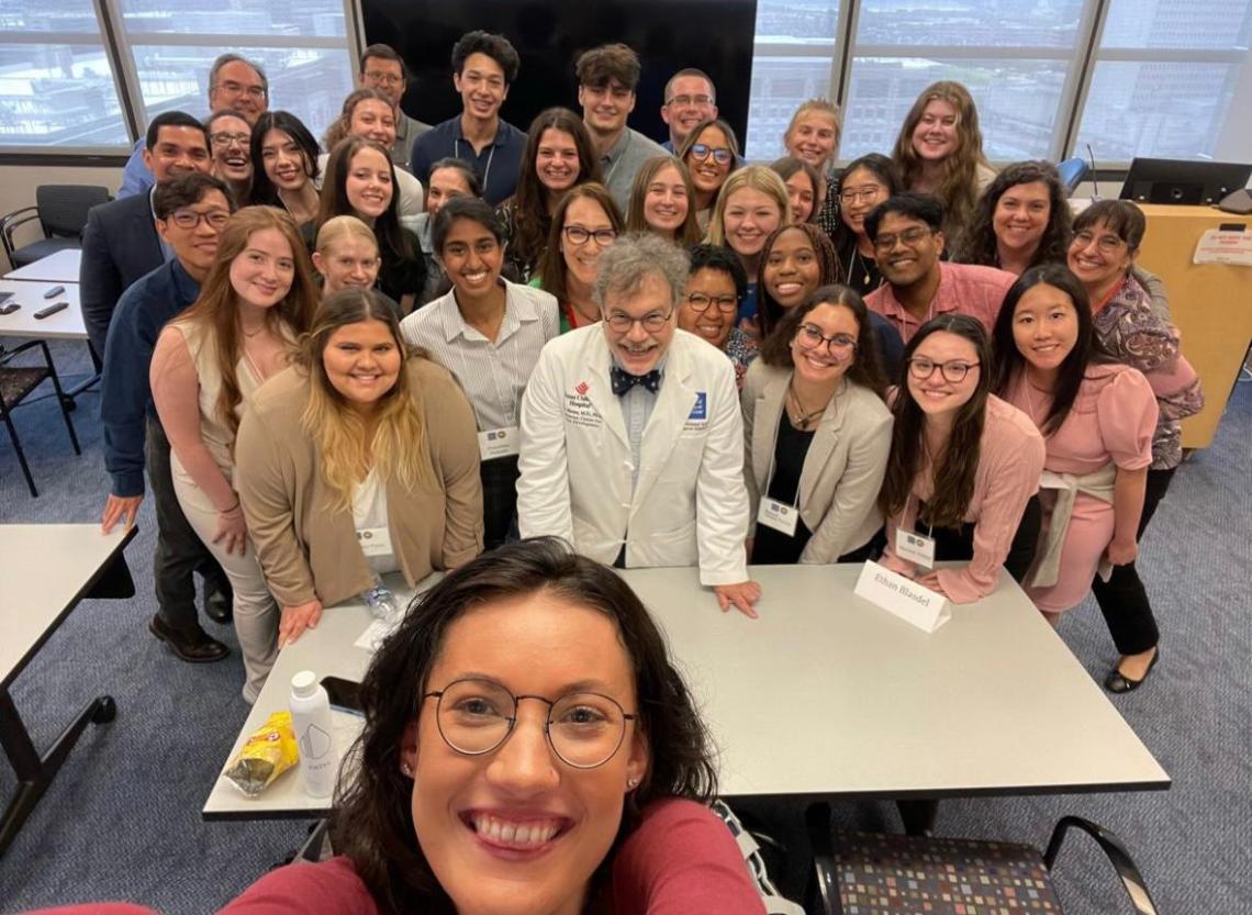 A smiling person taking a selfie with a large group of people posed shoulder-to-shoulder behind them.