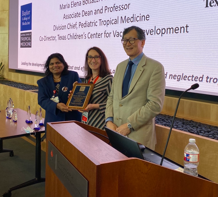 Dr. Sridevi Devaraj, Dr. Maria Elena Bottazzi and Dr. Jun Teruya stand in front of an auditorium. Dr. Bottazzi is holding a plaque.