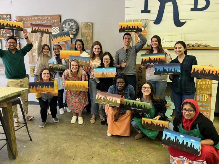 Pediatric Critical Care Fellowship members hold art they created during an event outside of the office. The art is a long painting showing the Houston skyline in a variety of backgrounds and atmospheres