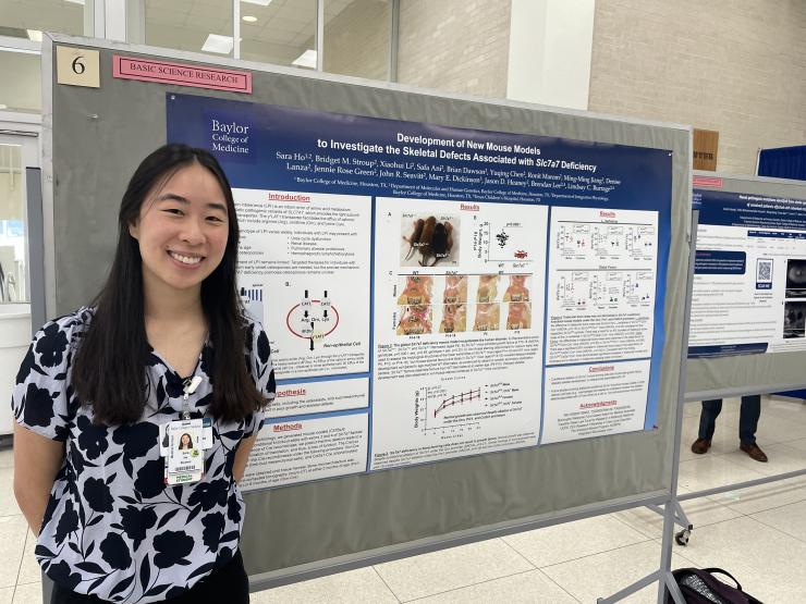 Sara Ho, wearing a black and white flowered top and her Baylor College of Medicine ID badge, stands in front of a poster showcasing her research