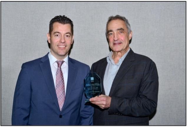 two men standing together smiling, with one holding an award plaque