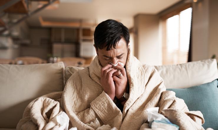 Man sitting on couch while wrapped in a blanket and blowing his nose