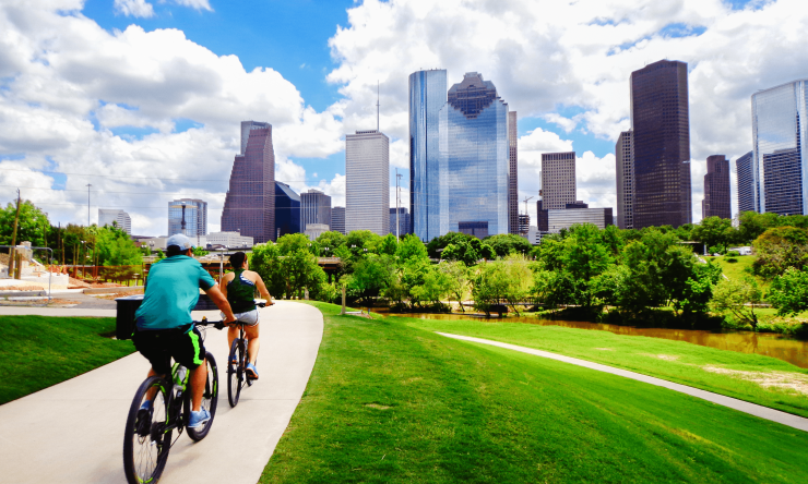 Two people ride bicycles toward downtown Houston, traveling across green grass with a blue, cloud-filled sky