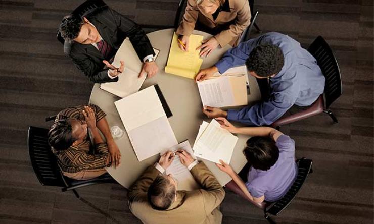 Six people seen from above sitting around a table examining papers