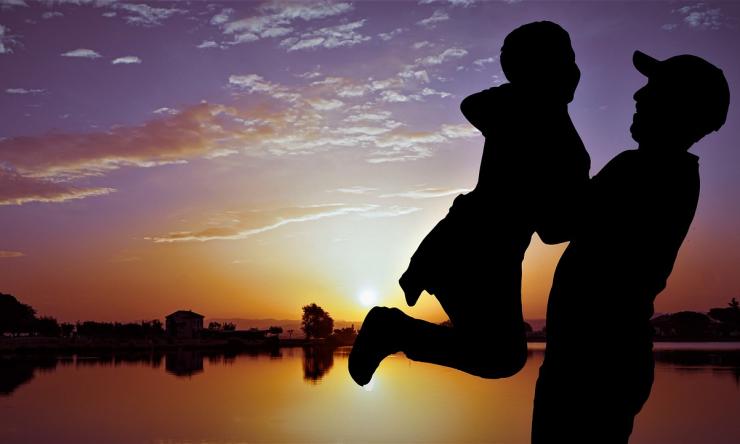 Photo of a sunset showing the silhouette of a father lifting his young son up in the air.