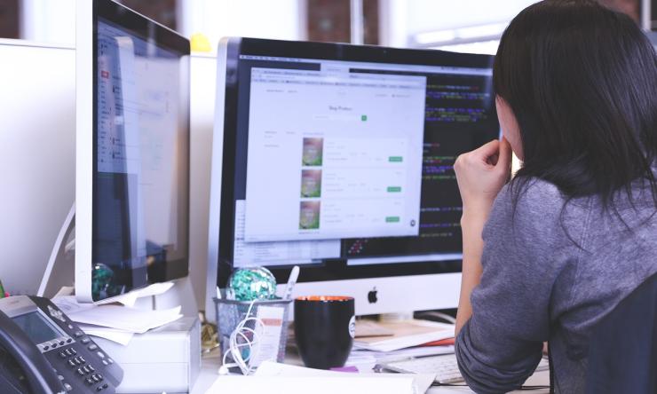 Photo taken from behind of a woman sitting at her desk computer hunched over  looking worried.