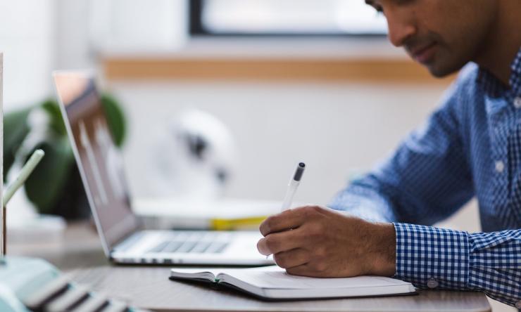 Photo of a man sitting at his desk, writing in a notebook while sitting next to his laptop